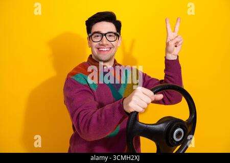 Ein lächelnder junger Mann posiert mit dem Lenkrad und macht eine Friedensgeste, die Charisma auf einem kräftigen gelben Hintergrund hervorhebt Stockfoto