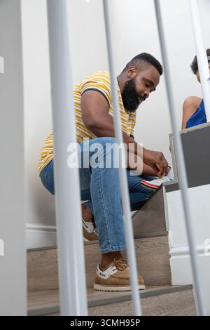 Afroamerikanischer Vater kniete auf einer Holztreppe durch weißes Geländer, schnürte Sneaker-Schnürsenkel für den Sohn Stockfoto