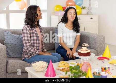 Vielfältige Mutter und Teenager-Tochter feiern 18. Geburtstag auf Wohnzimmer Sofa mit brennendem Kuchen Stockfoto