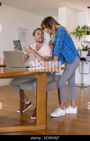 Paare arbeiten auf einem Tablet zusammen, während sie an einem hölzernen Esstisch mit Papieren für Kaffeetassen aus Laptops sitzen Stockfoto