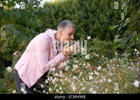 Reife Erwachsene Frau lehnt sich über Wildblumen im Garten des Hinterhofs und schröpft die Blüte neben dem Gesicht Stockfoto