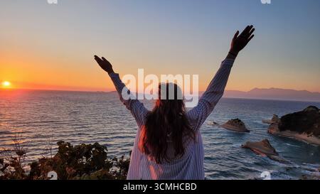 Eine Frau, deren Arme in Freiheit angehoben sind und den traumhaften Sonnenuntergang von den Klippen von Kap Drastis, Korfu Island, Griechenland, genießen Stockfoto