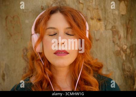 Rothaarige Frau, die Musik mit rosa Kopfhörern und geschlossenen Augen genießt, lehnt sich an einen Baumstamm Stockfoto