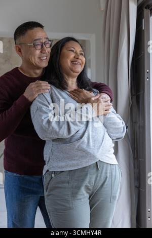 Diverse Paare umarmen sich, während sie am großen Fenster im Wohnzimmer mit Vorhängen stehen Stockfoto