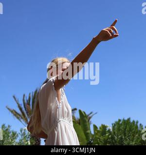 Eine schöne Frau in einem weißen Kleid zeigt ihren Finger zum klaren blauen Himmel und genießt den Sonnenschein Stockfoto