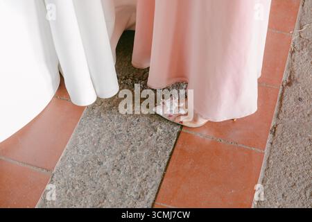 Eine Braut in einem rosafarbenen Kleid steht auf einem Steinboden und zeigt ihre zarten Schuhe bei einer Hochzeit im Freien. Stockfoto