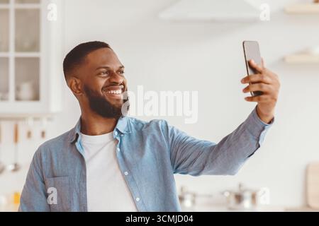 Zufriedener Tausendjähriger afroamerikanischer Mann mit Bart macht Foto am Telefon im Kücheninnenraum Stockfoto