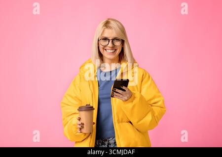 Positive Frau in gelber Jacke, die Kaffee zum Mitnehmen und SMS hält Stockfoto