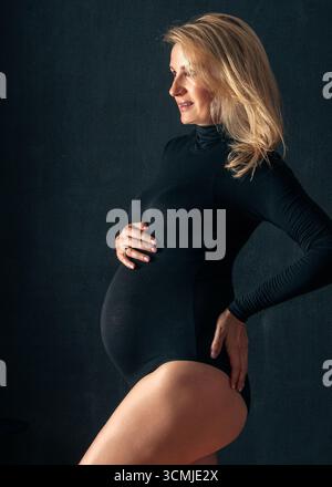 Schwangere Frau im schwarzen Body, die ihren Bauch berührt und lächelt. Moderne Mutterschaftsfotos über Mutterschaft, Schönheit und Lebensvorausschätzung Stockfoto