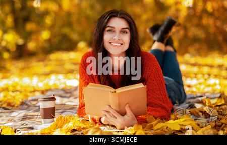 Lächelnde junge Frau mit Buch und Kaffee zum liegen gehen Auf Decke zwischen herbstlichen Blättern und warmen Herbst genießen Tag Stockfoto
