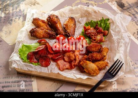 Appetitliche frittierte Hähnchenstangen, Speck, Peperoni-Würstchen und Nuggets mit frischem Salat und Kirschtomaten auf einem rustikalen Holzbrett. Stockfoto