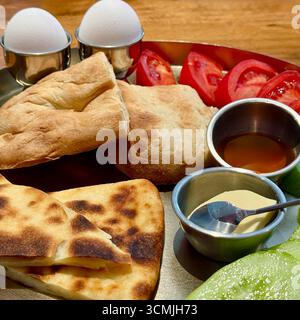 Nahaufnahme eines traditionellen georgianischen Frühstücks mit georgischem Imeruli Khachapuri, weich gekochten Eiern, Brot, Tomaten, Gurken, Butter und Honig Stockfoto