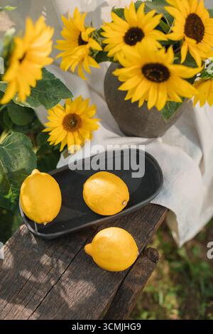 Nahaufnahme eines Strauchs Sonnenblumen (Helianthus annuus) in einer Vase mit frischen Zitronen auf einem Tablett auf einer Holzbank in einem Sommergarten Stockfoto