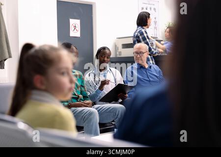 Porträt eines afroamerikanischen Arztes, der die Bedenken der Patienten über Nebenwirkungen von Medikamenten aufschreibt. Ärztin sitzt zwischen älterem Mann und jungen schwarzen Männern im klinischen Gesundheitsgespräch. Stockfoto