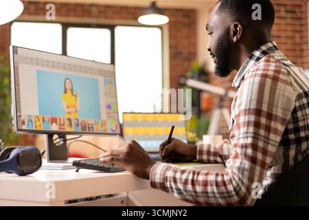 Professioneller Grafikeditor, der an einer Workstation mit zwei Bildschirmen Remote an der Bildretuschung arbeitet. Legere Kleidung, warmes Licht und die Innenausstattung aus Ziegelwänden spiegeln Fokus, Produktivität und modernen kreativen Prozess wider. Stockfoto