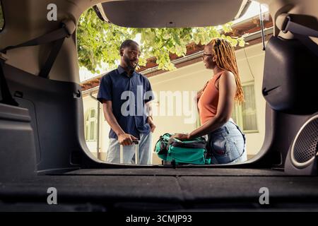 Schwarzer Mann und Frau, die Gepäck in das Auto laden und sich auf die Autofahrt vorbereiten, umgeben von Koffern und Sommersonne in der Auffahrt. Fröhliches Abenteuer mit Spaß und Freizeitreisen zusammen. Stockfoto