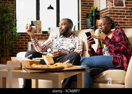 Glückliches schwarzes Paar entspannt sich auf der Couch in einer warmen, gemütlichen Wohnung und blättert auf Mobiltelefonen durch die sozialen Medien. Freund und Freundin genießen gute Zeit zusammen mit Snacks und Mahlzeiten zum Mitnehmen. Stockfoto