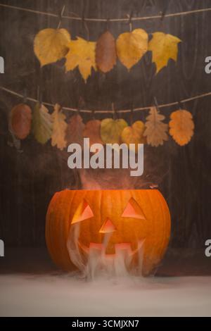Geschnitzter Halloween-Kürbis mit leuchtendem Gesicht gibt Rauch ab, der vor dem Hintergrund von lebhaften Herbstblättern das festliche Ambiente für die Saison unterstreicht Stockfoto