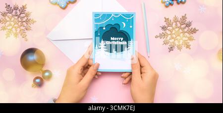 Weibliche Hände mit Grußkarte, Stift und Weihnachtsdekoration auf rosa Hintergrund, Draufsicht Stockfoto