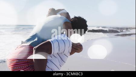 Mann, der eine Frau mit Huckepack am Sandstrand unter hellem Sonnenlicht barfuß trägt, mit sanften Wellen Stockfoto