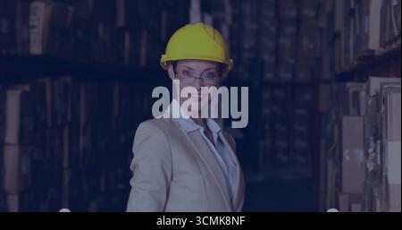 Frau mit Blazer, die Regale im Lagerhaus inspiziert, mit Kisten, Schutzhelm und Schutzbrille Stockfoto