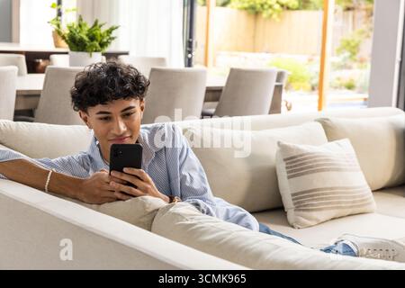 Mann, der sich auf einem cremefarbenen Sofa im Wohnzimmer zurücklehnt, auf dem Smartphone-Bildschirm scrollen, Kopierraum Stockfoto