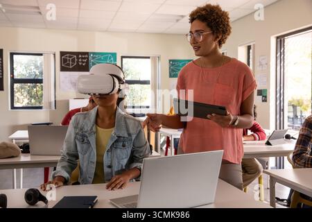 Lehrerin mit Tablet, die die chinesische Schülerin mit VR-Headset im Klassenzimmer mit Laptops begleitet Stockfoto