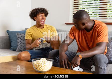 Verschiedene männliche Freunde sitzen auf einem Sofa im Wohnzimmer, spielen mit Controllern, Popcorn Stockfoto