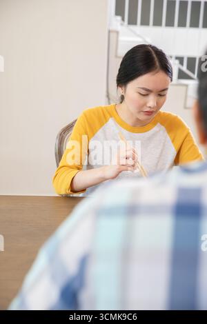 Asiatische Frau, die zu Hause am Esstisch sitzt und Essstäbchen mit dem Gast hält und sich auf das Essen konzentriert Stockfoto