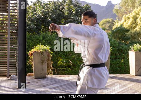 Ein Mann, der Kampfkunst-Punsch in Gi und Gürtel auf Holzdeck mit Hecken übt Stockfoto