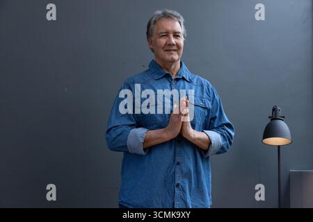 Senior Mann, der im Studio steht, trägt Denim-Hemd und dunkle Hosen bei Stehlampe Stockfoto