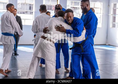 Vielfältige männliche Judoka feiert den Wettkampf, indem sie sich anschließt und in gis auf Matte mit Trophäe anfeuert Stockfoto