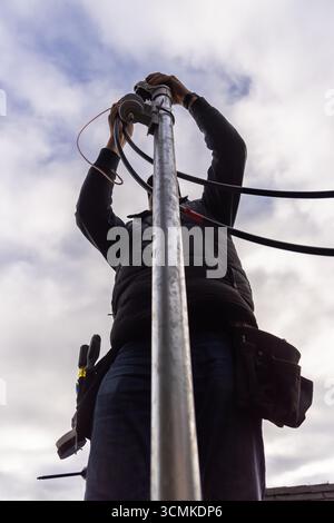 Telekommunikationstechniker, der in der Höhe arbeitet und Kabel an einem Metallmast mit bewölktem Himmel im Hintergrund installiert Stockfoto