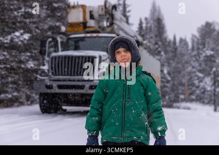 Kind, das bei Schneefall mit einem Schaufelwagen im Hintergrund in einem verschneiten Wald die Zunge ausstreckt Stockfoto