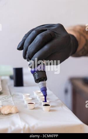 Tätowierer trägt schwarze Handschuhe, die violette Tinte in kleine weiße Tassen gießen und Farben für die Tätowierung vorbereiten Stockfoto
