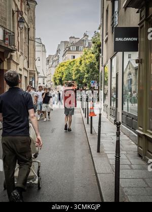 PARIS, FRANKREICH - 22. JUNI 2025: Ein Lieferarbeiter läuft durch eine enge Marais-Straße in Paris, mit einem Wagen zwischen Fußgängern und Ladenfronten, illus Stockfoto