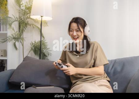 Eine junge Frau spielt zu Hause gern Videospiele, trägt Kopfhörer und hält einen Controller mit Spannung. Sie sitzt bequem auf einem Sofa und lächelt Stockfoto