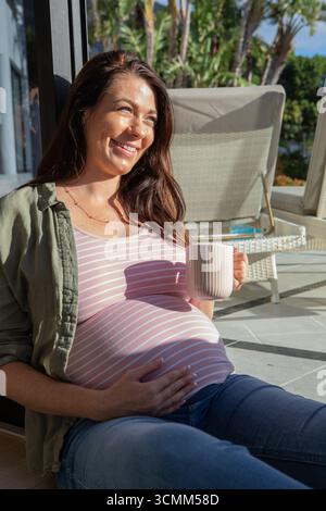 Schwangere Frau lehnt sich an eine Glastür und hält Kaffeetasse auf einer sonnendurchfluteten Terrasse mit Korbmöbeln Stockfoto