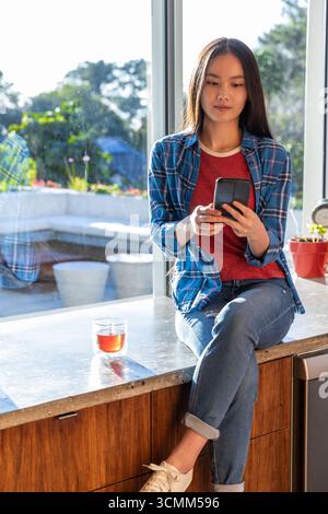 Asiatische Frau, die auf Marmortheke am Fenster sitzt und Smartphone mit Glasbecher hält Stockfoto