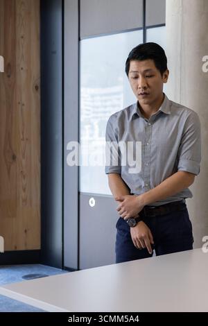 Asiatischer Mann, der hinter weißem Meeting-Tisch im modernen Büro steht und Armbanduhr überprüft Stockfoto