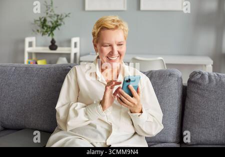 Fröhliche reife Frau, die Freude ausstrahlt, rollt durch ihr Handy und genießt einen ruhigen Moment. Stockfoto