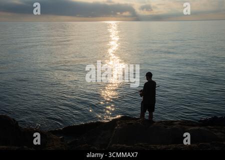 Silhouette eines jungen Mannes, der bei Sonnenaufgang auf dem Meer fischt. Fisherman, der Stab gegen aufgehende Sonne wirft. Friedliche Szene bei Sonnenaufgang an der Küste Stockfoto