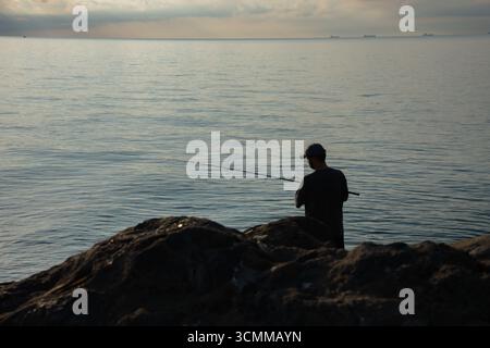 Silhouette eines jungen Mannes, der bei Sonnenaufgang auf dem Meer fischt. Fisherman, der Stab gegen aufgehende Sonne wirft. Friedliche Szene bei Sonnenaufgang an der Küste Stockfoto