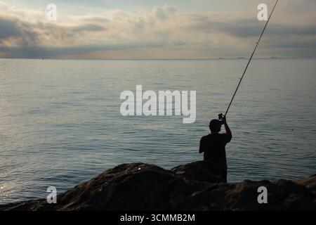 Silhouette eines jungen Mannes, der bei Sonnenaufgang auf dem Meer fischt. Fisherman, der Stab gegen aufgehende Sonne wirft. Friedliche Szene bei Sonnenaufgang an der Küste Stockfoto