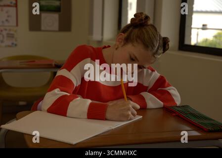 Mädchen in gestreiftem Pullover, das am Schultisch sitzt und in Notizbuch mit gelbem Bleistift schreibt Stockfoto