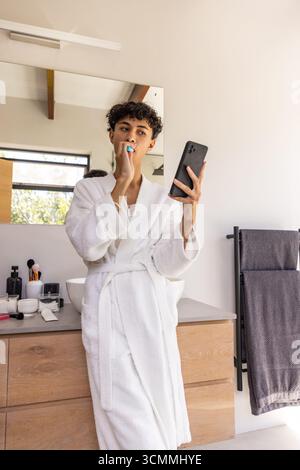 Mann Mitte 20 im Bademantel putzt Zähne mit blauer Zahnbürste im Badezimmer und hält Smartphone Stockfoto