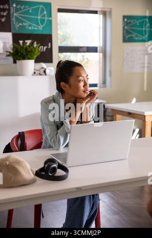Asiatische Studentin sitzt am Klassentisch mit Laptop mit schwarzen Kopfhörern und Baseballkappe Stockfoto
