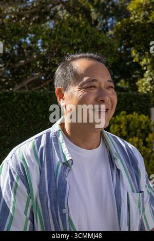 Reifer erwachsener asiatischer Mann, der im sonnendurchfluteten Garten steht und gestreiftes Hemd und T-Shirt trägt Stockfoto