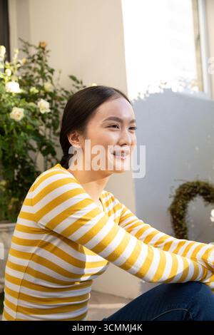 Asiatische Frau in ihren Zwanzigern, die auf dem Balkon sitzt, mit einer Rose, einem Kranz, Fensterläden, lächelnd Stockfoto