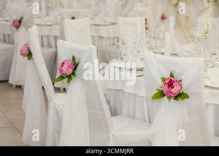 Wunderschöner Hochzeitsempfang mit weißen Stühlen mit weichem Stoff. Jeder Stuhl hat eine zarte rosa Pfingstrose und Grün für einen eleganten Touch Stockfoto